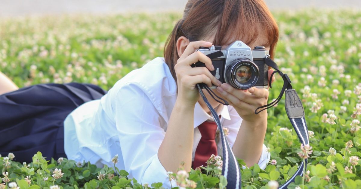 草原にうつ伏せてカメラを構える女子高生